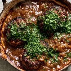 Close-up of thick seared hamburger steaks with dark brown caramelized edges in a glossy gravy with onion slivers.