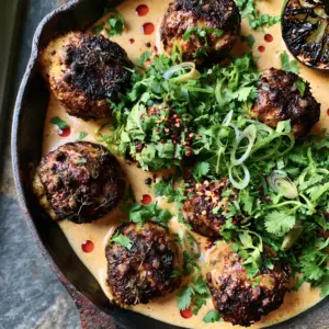 Close-up of seared jerk meatballs half-submerged in a pale-orange creamy curry sauce with red chili oil and scallions.
